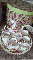 Photo of the full miniature tea set of Royal Doulton Country Rose pattern including teapot, creamer, sugar bowl, three cups, saucers, and serving plate, with round floral box in background.