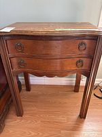 Front view of wooden side table showing two curved drawers with metal ring pulls and tapered legs on a light wood floor.