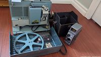 Full view of the vintage blue/beige Bell & Howell movie projector inside its carrying case with a film reel, next to the gray Bell & Howell Electric Eye movie camera and black carrying case.
