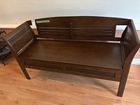 Front view of dark wood bench showing seat, backrest with horizontal slats, and solid legs on wood flooring.