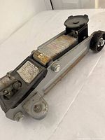 Hydraulic floor jack close-up showing main body, lifting pad, pump handle, and wheels with rust spots and instruction labels.