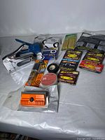Full lot overview showing Arrow staple tool with blue handle, multiple boxes of staples, utility blades, electrical tape rolls, Gorilla epoxy, and staple remover gauge on white background.