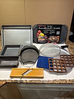 Full assortment of kitchen cookware and bakeware items laid on a kitchen counter including baking pans, Nordic Ware Pro-Cast grill/griddle in its box, splatter screen, mold trays, coasters, and slicer.