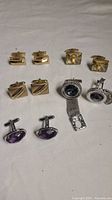 All 5 pairs of assorted metal cufflinks laid out on white surface, showing variety of styles and designs including gold-tone and silver-tone metal, some with stones and textured details.