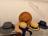 Photo of all five hats displayed together on a shelf showing the variety of brim sizes and colors including black and natural straw tones.