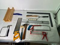 Full overview of all tools including wooden miter box, crosscut saw, coping saw, hacksaws, hammers, caulking gun, carpenter's square, and measuring tools arranged on flat surface.
