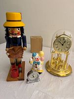 Full view of lot showing wooden nutcracker, porcelain clown figurine, gold-tone glass dome anniversary clock marked Mirado, silver-tone desk clock, and small box.