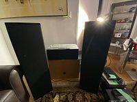 Two tall black bookshelf speakers with fabric grilles pictured upright against a sofa and wall.