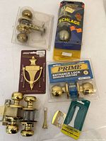 Overview photo showing various new door hardware including brass door knocker, entrance lock with key, brass door handles, Schlage knob and door stopper parts all laid out on white surface.