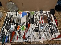 Wide view of miscellaneous kitchen utensils showing knives, scissors, graters, peelers, ladles, melon ballers, and other varied items arranged on a cloth.