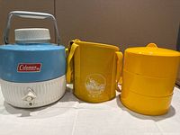 Full set showing a vintage blue and white Coleman coffee thermal on the left, a yellow insulated Kordco carry case in the center, and a yellow three-tier stackable food container on the right.