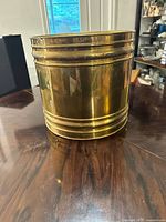 Front and side view of the large brass planter sitting on a wooden table, showing its shiny surface and raised horizontal bands.