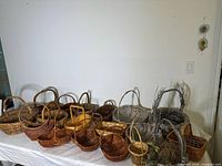 Photo showing 18 various baskets arranged on white table, displaying their shapes, handles, and woven textures including natural and assorted colors