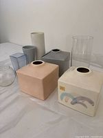 Eight assorted vases in multiple materials including ceramic, glass, and plastic with a variety of shapes and pastel/neutral colors