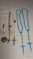 Photo showing six Catholic religious items including rosaries, necklaces, and medals arranged on a fabric surface. Items include blue plastic bead rosaries with crucifixes, white and red bead necklaces, metal medals, and pendants.