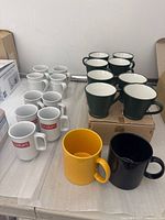 Photo showing 6 white Bodum logo mugs, 4 plain white mugs, 4 dark green mugs, and 2 extra large mugs (yellow and black) on top of cardboard boxes and table.
