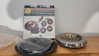 Photo showing two metal stove top cooker grills, one boxed and one used, along with a metal ring component, displayed on a wooden surface with the box showing usage instructions.