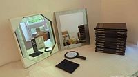 Two framed vanity mirrors: octagonal and rectangular shapes, and ten black handheld mirrors stacked, plus a black magnifying mirror and cleaning cloth on table.