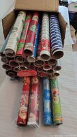 Box containing approximately 35 rolls of Christmas-themed wrapping paper in assorted colors and patterns.