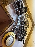 Wide view of various stainless steel kitchenware including bowls, plates, teapots, tumblers, and strainers arranged on white cloth on kitchen countertop.
