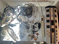 Photo of scattered power cords, extension cube, white 12-outlet power strip, and various AV cables including HDMI and component cords