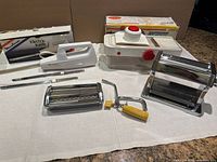 Front view of three kitchen appliances on white surface including Moulinex electric knife, Starfrit Supreme slicer and Grazia pasta maker with original boxes in background.