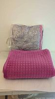 Two blankets folded, one in clear bag showing grey quilted blanket with pink trim, other solid pink blanket in front.