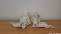 Four white porcelain cherub angel figurines grouped on a wooden surface, showing variety of poses and wing details, with minor surface wear visible.