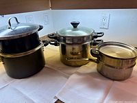 Photo showing four pots: two black-coated pots stacked on the left, one stainless steel stockpot with glass lid with steamer insert inside on the right, and another stainless steel pot/insert in front.