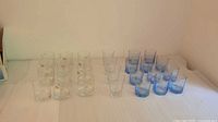 Overview of multiple clear and blue glass cups arranged on a flat surface, includes short tumblers with gold decoration, medium clear glasses, and blue glasses.