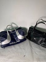 Three vintage Adidas duffel bags set on white cloth. Navy and silver bag with large logo on left, two black bags on right.