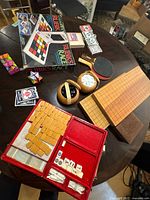 Eclectic tabletop games lot displaying Rubik’s Race, Go board with stones in wooden bowls, red Mahjong set with tiles and scoring sticks, table tennis paddles, decks of playing cards, and foldable snake puzzles on a dark wood round table.