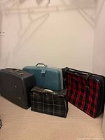 Four vintage suitcases grouped in a corner: black hard-shell, teal hard-shell, red and black plaid soft-sided, green and black plaid soft-sided Qantas bag