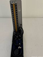 Mercurial sphygmomanometer open showing vertical mercury measurement scale, inflation bulb and coiled tubing inside textured gray metal case.