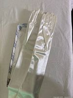 Photo showing the gloves laid on a flat surface inside the original box. The gloves are made from shiny white satin, extending above the elbow with a smooth finish.