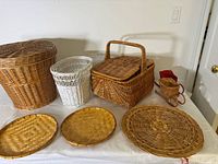 Seven woven and wicker items displayed on white tablecloth including large lidded basket, white waste basket, two round trays, two oval trays, miniature sled basket, and small red basket.