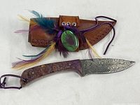 Full view of the hunting knife with Damascus steel blade and purple wood handle alongside the brown leather sheath decorated with colorful feathers and a green stone metal tag.