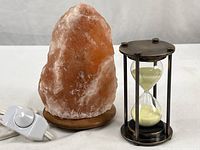 Himalayan salt lamp with wooden base next to steel hourglass with white sand on a white background, lamp cord with inline dimmer visible.