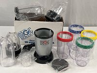 Full set of Magic Bullet components displayed, showing base unit, four mugs with colored lids, and accessories, on a white surface with original box.