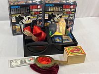 Two sealed Merlin's Magic Show kits with contents displayed in black trays, and one wooden box with assorted magic props arranged in front.