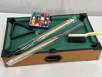Full view of tabletop pool table showing the green felt playing surface, black corner pockets, wooden finish, two pool cues in plastic wrapping, set of billiard balls in box, a chalk cube, rack, and a brush.