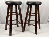 Two 26-inch height wooden bar stools with black leather round padded seats, sturdy legs, and connecting rungs.