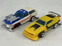 Two vintage Lionel plastic slot cars shown side by side on white background. Blue and white car with large engine detail and stars on roof. Yellow car with rear spoiler missing front tires.