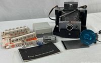 Polaroid 100 Land Camera with its accessories including flash attachment, clear flashbulbs, instructions booklet, portrait attachment case cover, and Canon battery charger.