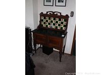 Full front view of the English wooden wash stand featuring a dark stone top, two cabinet doors, and a tile backsplash with floral and green tiles.