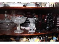 Shelf display showing clear pressed glass pedestal bowls and candy dish, along with some cranberry glass pitchers and bowls in the background