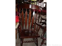 Front view of one solid wood rocking chair showing the curved backrest with vertical slats and turned spindle arms and legs.
