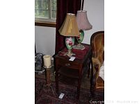 Two antique lamps with floral painted glass bodies and fabric shades on a dark wood end table with drawer and shelves.