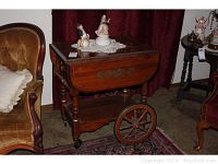 Photo of full wood tea cart with drop-leaf sides, large wood-spoked wheels, and a crocheted doily on top holding porcelain figurines.