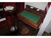 Photo showing the antique school master's desk with green felt surface, the tall plant stand with two shelves and lace doilies, and the carved wooden stool beneath the desk.